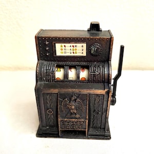May include: A vintage-style metal slot machine bank with a dark bronze finish. The front features an embossed eagle emblem above the text GOLDEN EAGLE. It includes a side lever, three spinning reels with fruit symbols, and a payout chart at the top.