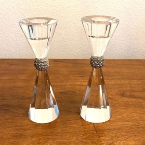 May include: A pair of clear glass candle holders with a crystal-like design. Each holder has a round, crystal-like embellishment around the middle.
