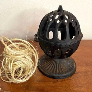May include: A black cast iron yarn ball holder with a decorative design. The holder is on a wooden surface next to a ball of twine.