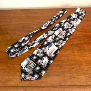 May include: A black necktie with a repeating pattern of computers, monitors, and the words "e-mail" and "internet" in white and blue. The tie is made of a smooth fabric and is displayed on a wooden surface.