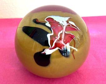 May include: A spherical glass paperweight with a smooth, polished surface. The base color is a muted olive green, featuring internal abstract swirls of deep black, vibrant red, and thin, opaque white glass filaments throughout.