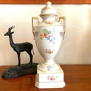 May include: A white porcelain vase with a floral design and gold accents. The vase has a lid and two handles. The vase is on a square base.
