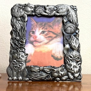 May include: A silver-toned picture frame with a cat design. The frame features a raised design of cats playing with yarn balls and bows. The frame is designed to hold a 4x6 inch photo.
