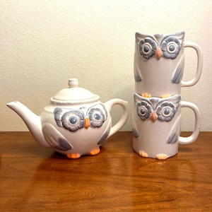 May include: A white ceramic teapot and two mugs with owl designs. The teapot and mugs have blue and gray owl faces with orange feet.