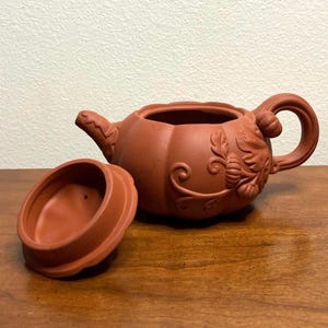 May include: A terracotta teapot shaped like a pumpkin with a floral design. The teapot has a lid that is separate from the teapot.