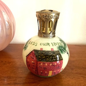 May include: A white ceramic oil burner with a gold metal lid. The burner has a red brick house with a green roof and the words "Bless Our Home" painted on it.