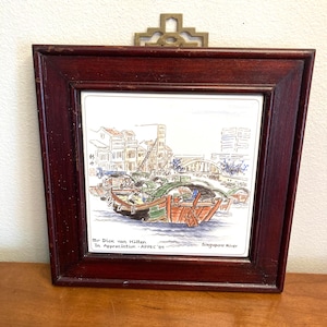 May include: A framed tile with a hand-drawn illustration of boats on the Singapore River. The tile is white with a brown wooden frame. The text on the tile reads "Mr Dick van Hilen In Appreciation - ADPEC '81 Singapore River".