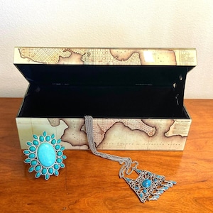 May include: An open wooden jewelry box with a map design on the lid. The box is empty, but there is a turquoise necklace and a turquoise ring on the table next to it.