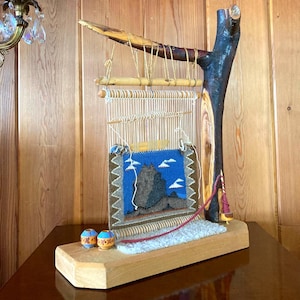 May include: A wooden loom with a blue and brown woven tapestry depicting a mountain scene. The loom is made of natural branches and has a wooden base. Two small, colorful, round beads are on the base.