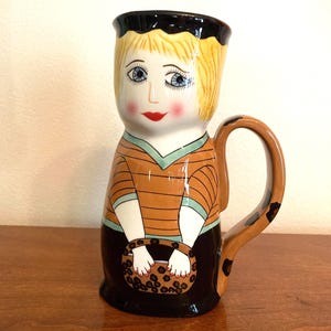 May include: A ceramic mug in the shape of a woman, featuring a blonde hairstyle and a black hat. The mug has a brown and orange striped shirt, a black skirt, and a handle with a leopard print design. The woman is holding a basket.