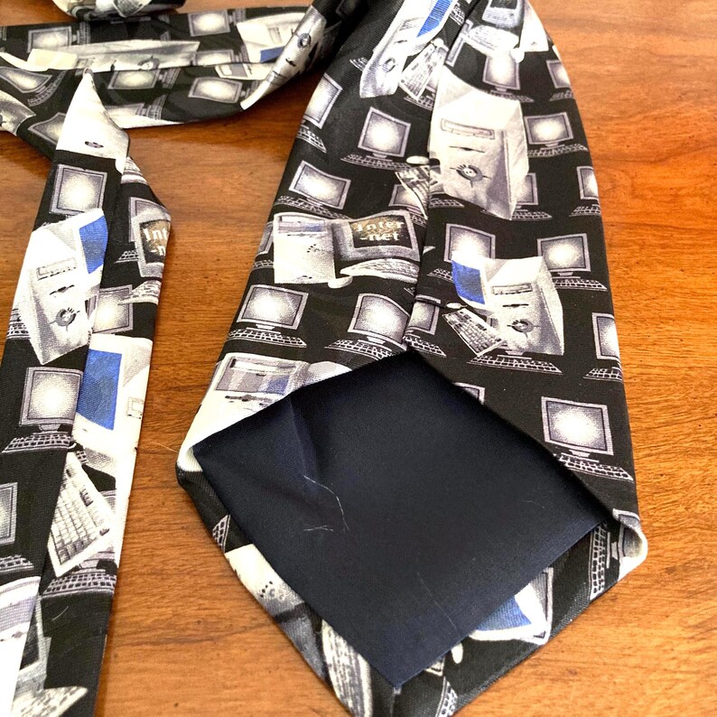 May include: A black necktie with a repeating pattern of vintage computer monitors, keyboards, and the word "Internet". The tie has a blue and white accent color. The tie is made of a smooth fabric.