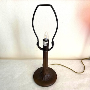 May include: A table lamp base featuring a brown, textured stem resembling a tree trunk with a circular base. The lamp includes a black metal harp for shade attachment and a single light bulb socket with a clear, pointed LED bulb installed.