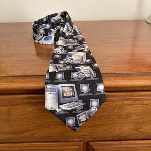 May include: A black necktie featuring a repeating pattern of vintage computer monitors, keyboards, and computer towers in shades of white and gray. The tie also has the words "e-mail" and "internet" printed on it.