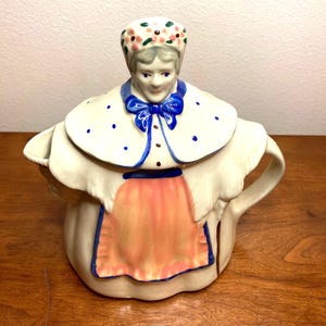 May include: A ceramic teapot shaped like a woman wearing a white dress with blue and pink accents. The teapot has a floral headband, a blue bow, and a white collar with blue polka dots.