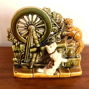 May include: A ceramic planter shaped like a spinning wheel with a cat and dog figurine. The spinning wheel is green and brown with a white dog and orange cat figurine. The planter has the text "K & S" on the bottom.