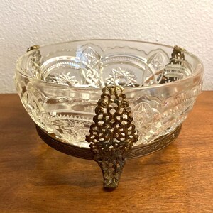 May include: A clear glass bowl with a cut glass design sits on a gold metal stand with three legs. The stand has a decorative, openwork design.