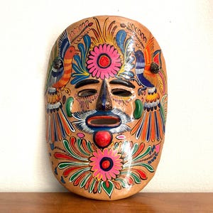 May include: Hand-painted, oval-shaped mask with a tan base. The mask features a stylized face with a blue nose, a red mouth, and a white mustache. It is decorated with colorful birds, flowers, and intricate patterns in shades of pink, orange, blue, and green.