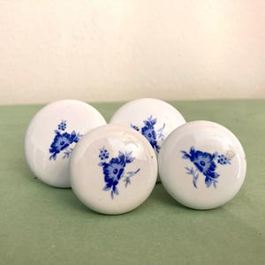 May include: Four white ceramic cabinet knobs with blue floral designs. The knobs are round and have a glossy finish. The blue floral pattern is a delicate, detailed design. The knobs are arranged on a green surface.