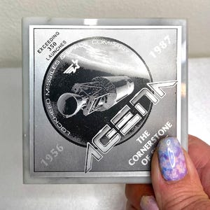 May include: Square marble coaster with a silver metallic design featuring the Lockheed Missiles & Space Company logo, the word "AGEAN", and the text "The Cornerstone of". Includes the years 1956 and 1987.