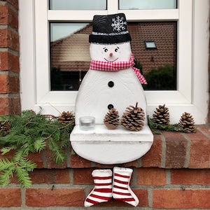 Könnte beinhalten: Weißer Holzschneemann mit einem schwarzen Hut und einem rot-weiß karierten Schal. Der Schneemann hat eine kleine schwarze Knopfnase und eine schwarze Schneeflocke auf seinem Hut. Der Schneemann sitzt auf einem weißen Regal mit einem kleinen Glas-Kerzenhalter. Der Schneemann trägt rot-weiß gestreifte Stiefel.