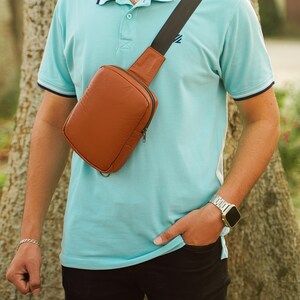 Puede incluir: Un bolso bandolera de cuero marrón con una correa ajustable negra. El bolso tiene una cremallera y una solapa con cierre magnético. El bolso lo lleva una persona que lleva una camisa polo azul claro y pantalones negros.