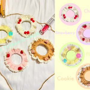 May include: Five crocheted hair scrunchies with different designs. The scrunchies are in the colors white, yellow, pink, brown, and beige. The designs include cherries, strawberries, flowers, and cookies. The scrunchies are labeled "Cherry", "Strawberry", "Flower", "Cookie", and "Flower".