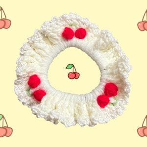 May include: White crocheted hair scrunchie with red cherry embellishments. The scrunchie has a ruffled edge and is decorated with five cherries.