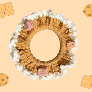 May include: A brown crocheted hair scrunchie with white trim and various food-themed charms, including a cookie, a cake, and a cartoon animal.