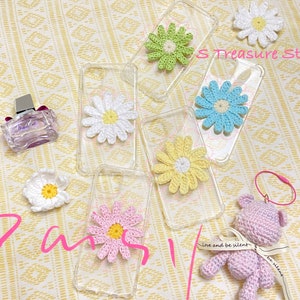 May include: Six clear phone cases with crocheted flower designs. The flowers are in various colours, including white, light blue, light green, and pink. The cases are on a patterned surface. The text "S Treasure Studio" is visible in the image.