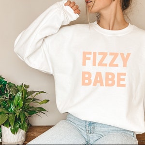 May include: White sweatshirt with the text "FIZZY BABE" in peach.