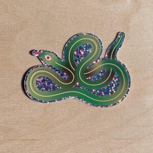 May include: A die-cut sticker featuring a coiled green snake with a glittery holographic finish. The snake has a smooth, dark green body with lighter green stripes and a small red eye. The background of the sticker is filled with a dense, sparkling glitter texture that reflects various colours.