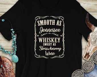 Può includere: Maglietta nera con un'etichetta in stile vintage che recita "Smooth as Jennessee Whiskey Sweet as Strawberry Wine". La maglietta è appoggiata su una superficie di legno con un paio di stivali da cowboy marroni, una collana turchese, una fibbia di cintura argentata e un teschio di mucca.