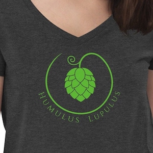 Può includere: Una maglietta a collo a V di colore grigio scuro con una grafica verde di un cono di luppolo in un cerchio con il testo "Humulus Lupulus".