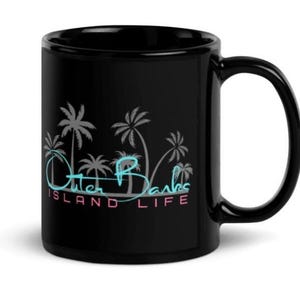 Puede incluir: Taza de cerámica negra con asa curva. La taza presenta un diseño con palmeras y las palabras "Outer Banks ISLAND LIFE" en una fuente de estilo neón, con colores azul, rosa y blanco.