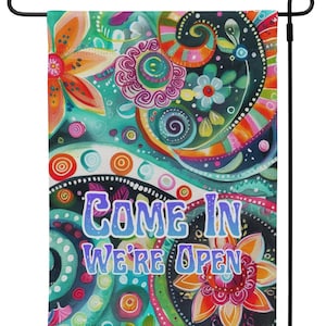 May include: A colorful garden flag with a vibrant floral design and the words "COME IN WE'RE OPEN" in blue and purple. The flag features a mix of orange, pink, green, and teal hues, perfect for outdoor decor.