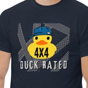 Può includere: Una t-shirt blu navy con un anatroccolo di gomma giallo che indossa un berretto da baseball blu. L'anatroccolo ha il testo "4x4" sul petto e il testo "DUCK RATED" sotto. Lo sfondo è un motivo grigio di battistrada di pneumatici.