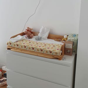 May include: A wooden baby changing table with a patterned changing mat featuring koala bears. A teddy bear, toys, and baby supplies are on the changing table, which is placed on top of a white chest of drawers.