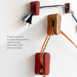Könnte beinhalten: Kabelorganisatoren aus Holz in verschiedenen Formen und Farben sind an einer weißen Wand montiert. Die Organisatoren halten Kabel und Seile. Der Text auf dem Bild lautet: "Elegant servants as cable management, cable routing, wall cable holder and rope holder."