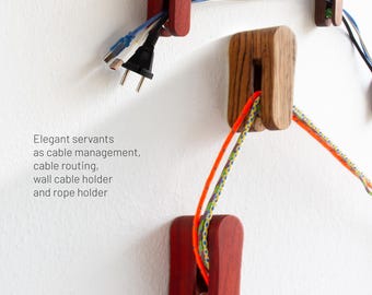 May include: Wooden cable organisers in various shapes and colours are mounted on a white wall. The organisers are holding cables and ropes. The text on the image says "Elegant servants as cable management, cable routing, wall cable holder and rope holder."