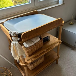 Op de afbeelding: Een houten commode met drie planken. De bovenste plank heeft een lichtblauwe commodemat. De middelste plank heeft een rieten mand gevuld met witte katoenen luiers. De onderste plank heeft een rieten mand. Een knuffelkoala hangt aan de zijkant van de commode.