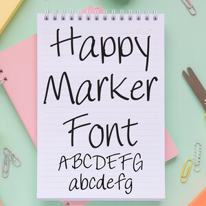 May include: A spiral-bound notepad with the words "Happy Marker Font" written in black marker. The alphabet is displayed in both uppercase and lowercase letters. The notepad is on a light green surface with pink and yellow stationery.