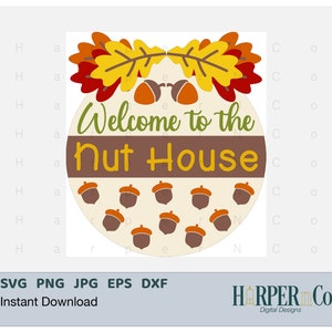 May include: A round, cream-colored sign with the text "Welcome to the Nut House" in brown, green, and yellow lettering. The sign is decorated with two autumn leaves in red, orange, and yellow, and a border of brown acorns.
