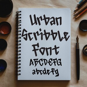 May include: A white spiral-bound notebook features the words "Urban Scribble Font" in black, bold lettering. The alphabet is shown in uppercase and lowercase below. Ink pots, pencils, and a pen are also visible.