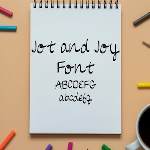 May include: A white spiral-bound notepad with the words "Jot and Joy Font" written in black script. The alphabet is displayed in both uppercase and lowercase. Colored markers and a coffee cup surround the notepad.