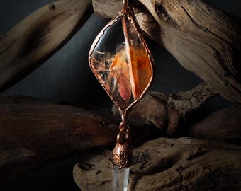 May include: A copper-colored pendant necklace with a clear, leaf-shaped center containing orange and white inclusions. A small, clear crystal hangs from the bottom. The necklace is displayed against a backdrop of weathered wood.