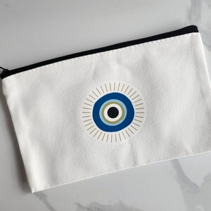 May include: White canvas pouch with a black zip and trim. The pouch features a circular design of a blue eye with gold and black accents. The pouch is rectangular and appears to be made of a sturdy material.