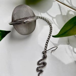 May include: A stainless steel tea infuser with a chain and a decorative snake charm. The infuser is a mesh ball with a hinged closure. The snake charm is detailed and hangs from a chain. The background is white with green leaves.