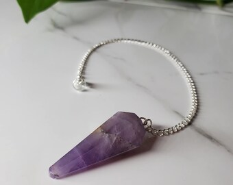 Pode incluir: Um pêndulo de cristal de ametista roxo em uma corrente prateada. O pêndulo é pontudo e tem uma textura natural e áspera.