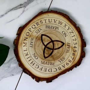 May include: Wooden Ouija board with a circular shape and a brown wood grain finish. The board features a black triquetra symbol in the center, surrounded by letters, numbers, and the words "Yes", "No", "Maybe", and "Wait".