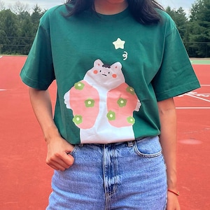 Puede incluir: Una camiseta verde con un animal de dibujos animados blanco que lleva una chaqueta estampada en rosa y verde. El animal tiene una estrella sobre su cabeza.